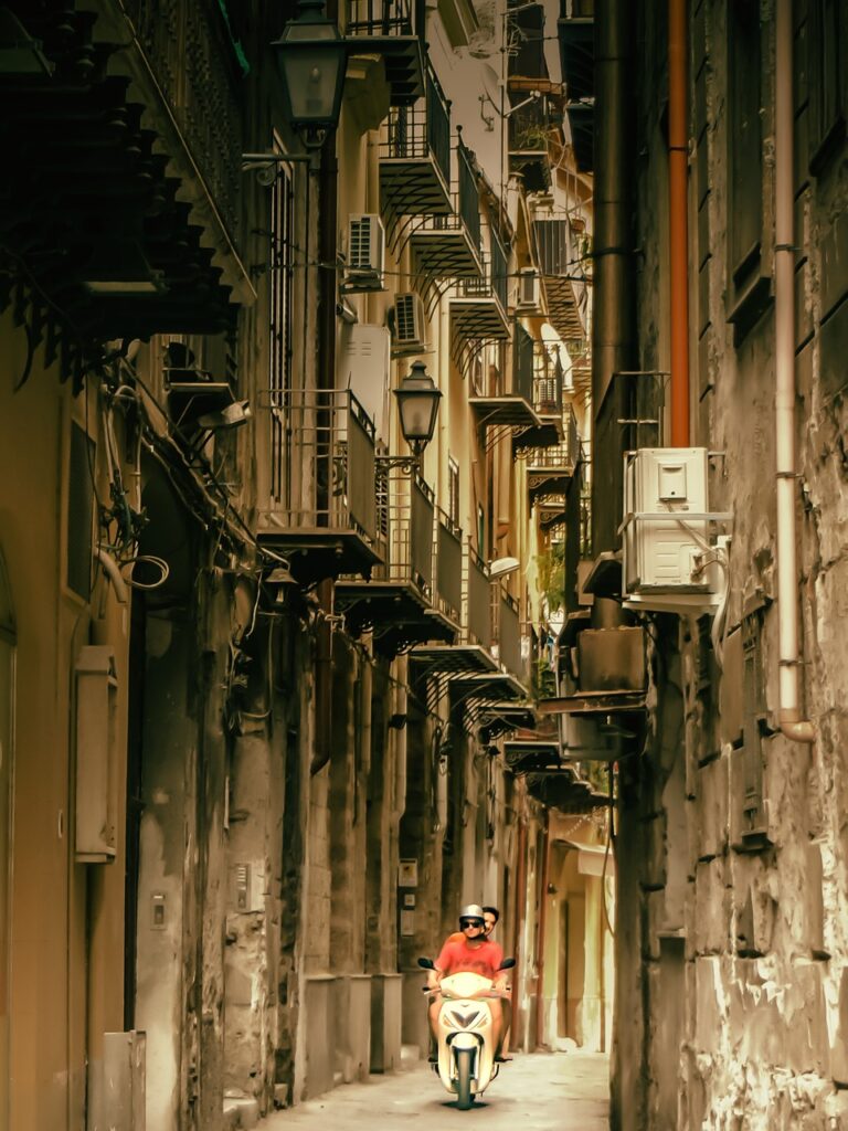 Two men driving a vespa down a narrow palermo street with balconies in the background.